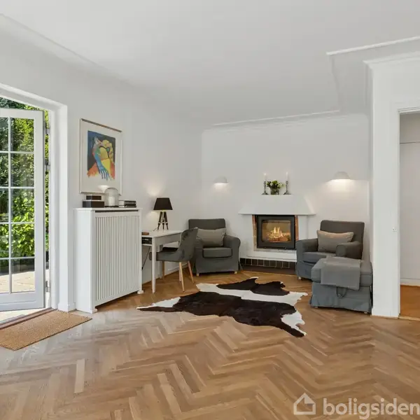 Stue med to komfortable lænestole ved siden af en moderne pejs, på et trægulv med en sort-hvidt tæppe. Store vinduer åbner ud til en terrasse med grøn udsigt.