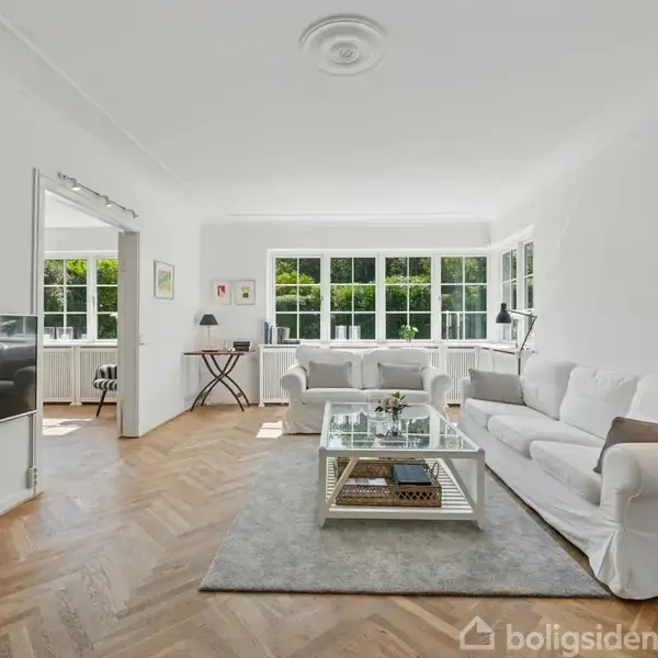 En stue med en hvid sofa og et glas sofabord. Rummet har trægulv, store vinduer der giver naturligt lys, og en vægmonteret TV. En lampe står ved sofaen.