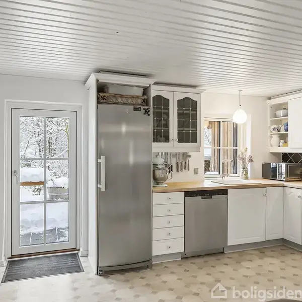 Et køkken med hvide skabe og stål køleskab står ved siden af en dør, der fører ud til et snedækket område. Et rundt lys hænger over træbordpladen.