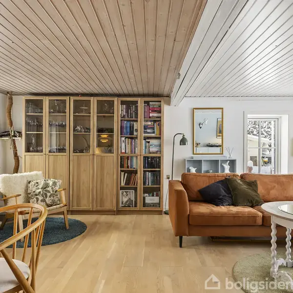 En stue med en brun sofa foran en væg med bogreoler og glaslåger. Et rundt sofabord står foran sofaen. Rummet har lyst trægulv og loft.