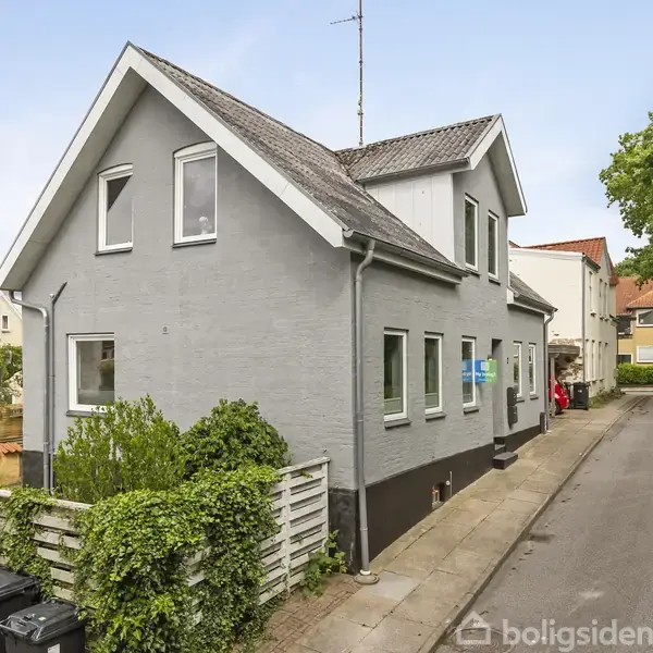 Et gråt, to-etagers hus står stille på en vej omgivet af grønne buske og træer. Flere skraldespande er placeret foran en hvidmalet hegn langs huset.