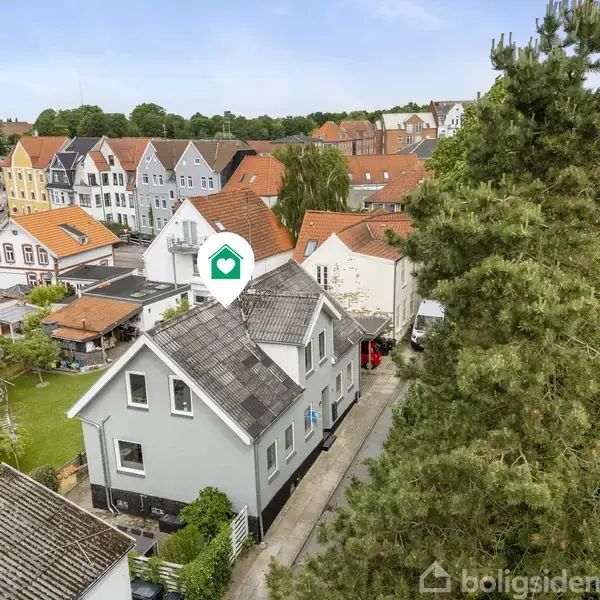 Et hvidt hus med mørkt tag ses fra en fugleperspektiv. Intet sker i billedet. Omgivet af andre huse og træer i et boligområde.