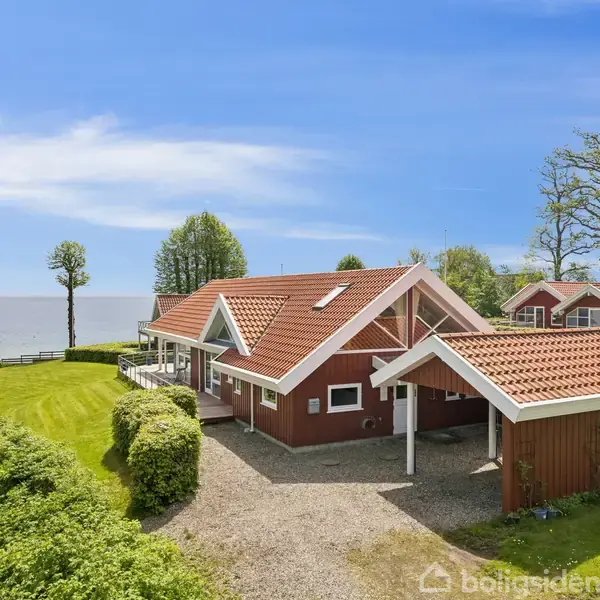 Rødt hus med terrasse og carport står ved vandet i grøn have omkranset af buske og græsplæner. Blå himmel og træer i baggrunden flankerer udsigten til havet.