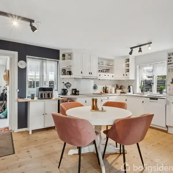 Et rundt bord med fire lyserøde stole står i et lyst køkken. Køkkenet har hvide skabe, træbordplade, og en opvaskemaskine. Et køleskab er dækket med magneter og sedler.