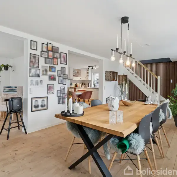 Spisebord med stole står i en rummelig stue. Væggen har mange billedrammer. Der er en trappe og store planter i baggrunden, og et sofahjørne i venstre side.