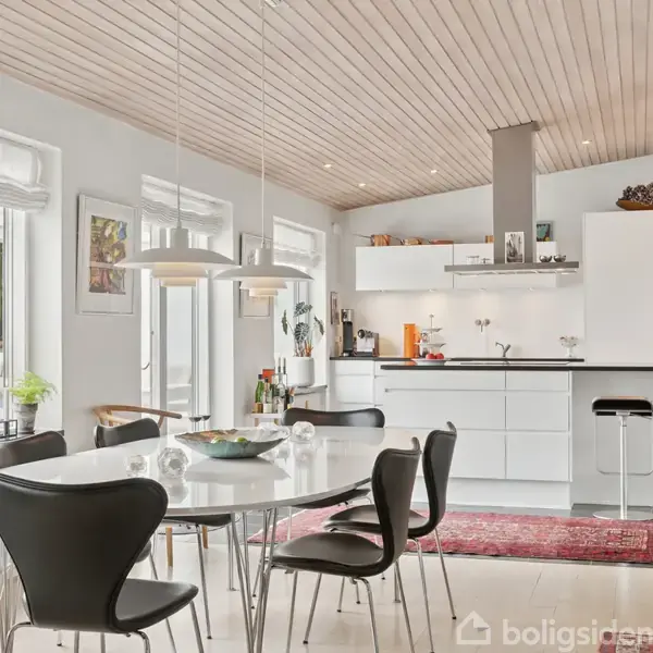 Et moderne køkken med hvidt interiør, spisebord og sorte stole i forgrunden. Køkkenet har træloft, store vinduer og flere planter og pyntegenstande. En rød løber ligger på gulvet.