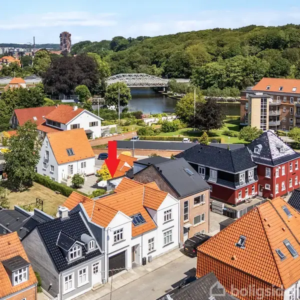 Hus markeret med en rød pil står blandt huse med røde tage i et beboelsesområde. En flod samt bro ses i baggrunden, omgivet af træer og lejlighedskomplekser.