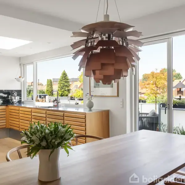 Loftlampe hænger over et spisebord i et moderne køkken med træelementer. I det lyse rum ses en buket blomster og udsigt over et boligområde gennem store vinduer.