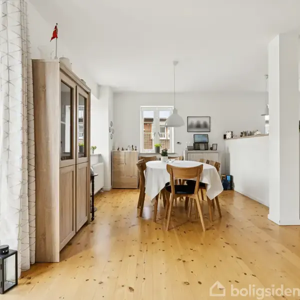 Spisebord står i en lys stue med træmøbler og trægulv. Et vitrineskab og loftlampe omgiver bordet med hvid dug. Køkken ses i baggrunden.