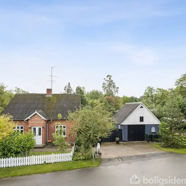 Rødt murstenshus med sort tag står bag en lille hvid stakit. Til højre en garage med sadeltag. Omgivet af grønne træer og en indkørsel med parkeret bil.