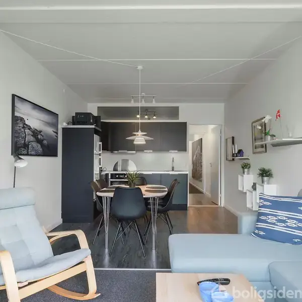 Stue med grå sofa og gyngestol til venstre, opholdsområde med spisebord og stole i midten, køkken bagerst. Væggene har hylder og billeder, der hænger ved siden af lampene.