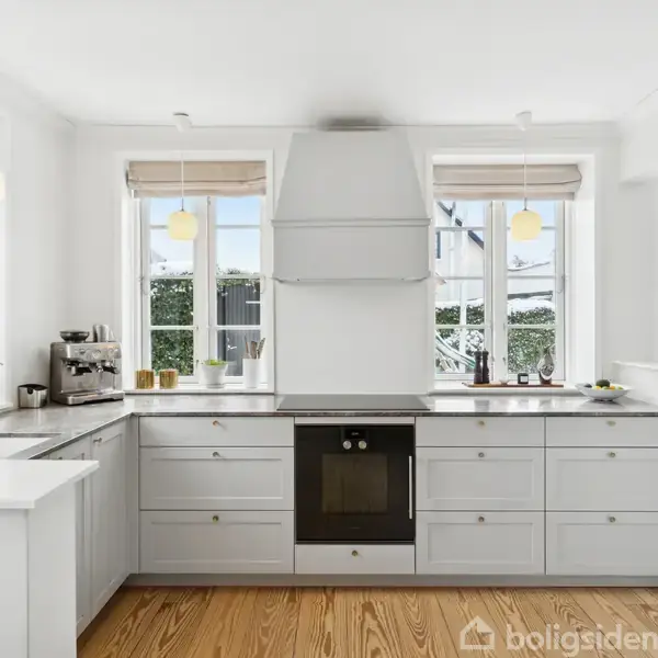 Et hvidt køkken med trægulv, udstyret med kogeplade og ovn i midten, omgivet af skabsplads. Store vinduer lader naturligt lys strømme ind.