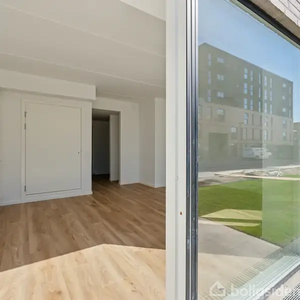 En lys stue med trægulv set indefra en åben glasskydedør. Minimalistisk køkken indbygget til venstre. Udenfor ses en græsplæne og moderne lejlighedsbygninger.