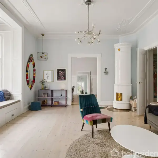 Stue med moderne indretning; en pejs er midtpunktet. En sofa og stol står rundt et ovalt sofabord. Store vinduer giver lys, og farverigt kunstværk pynter væggene.
