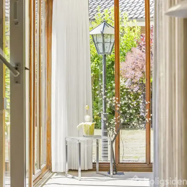 En gennemsigtig glasbord med en hvid vase står i et lyst rum fyldt med sollys. Udenfor ses grønne planter og et tag, der skaber en fredelig stemning.