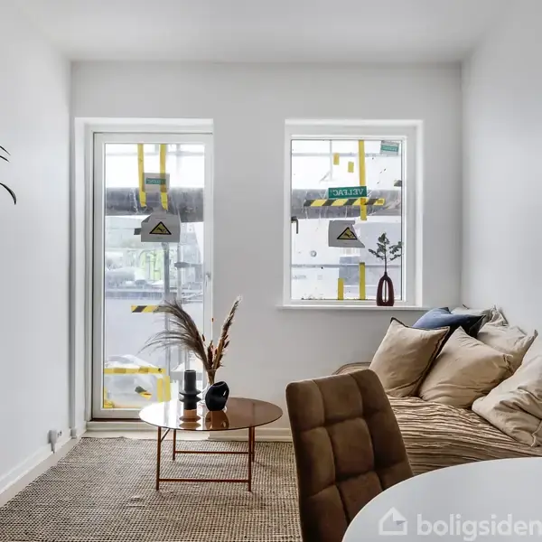 Stue med sofa fyldt med beige puder, placeret ved siden af bord med vaser. Store glasdøre og vinduer med opmærkning åbner mod en altan. Plante står i hjørnet.