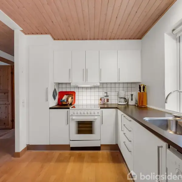 Et hvidt køkken med ovn og komfur i midten, omgivet af skabe. En vask med kromarmatur er på højre side nær et vindue med udsigt til en have.