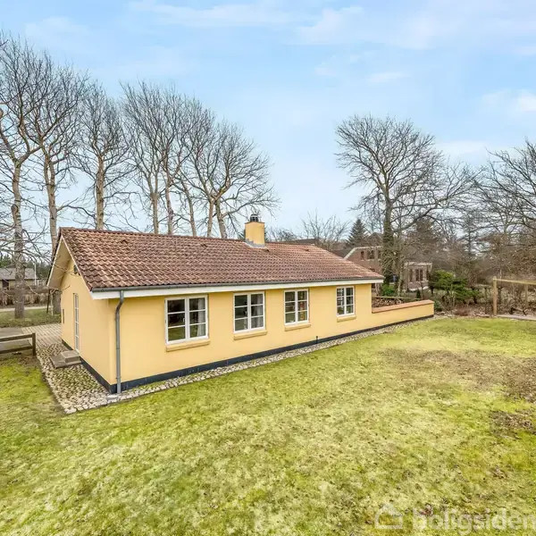 Et gult hus med rødt tag står stille omgivet af et frodigt grønt græsareal. Træer omkranser ejendommen i en rolig, skovlignende baggrund.