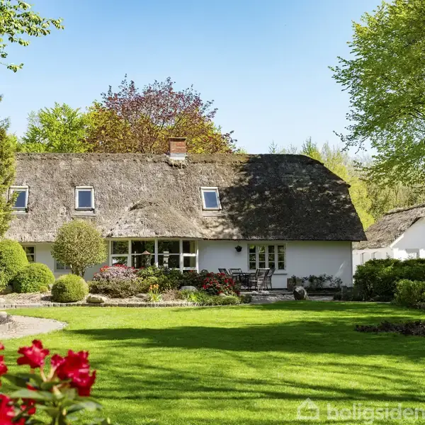 Et hvidt bindingsværkshus med stråtag ligger forankret i en frodig have med blomsterbede og veltrimmede buske. Sollys skinner over den grønne græsplæne og omgivende træer.