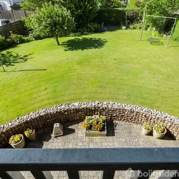 En græsplæne med gyngestativer står i en baghave omgivet af hække. Foran er en brostensbelagt terrasse med blomsterkrukker og en blomsterkasse, omkranset af en stenmur.