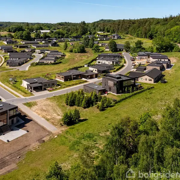 Boligkvarter med moderne huse langs snoede veje i grønt landskab. Omkranset af skov og åbne marker, skaber en fredelig forstadsstemning. Husene er adskilt af velplejede græsplæner og træer.