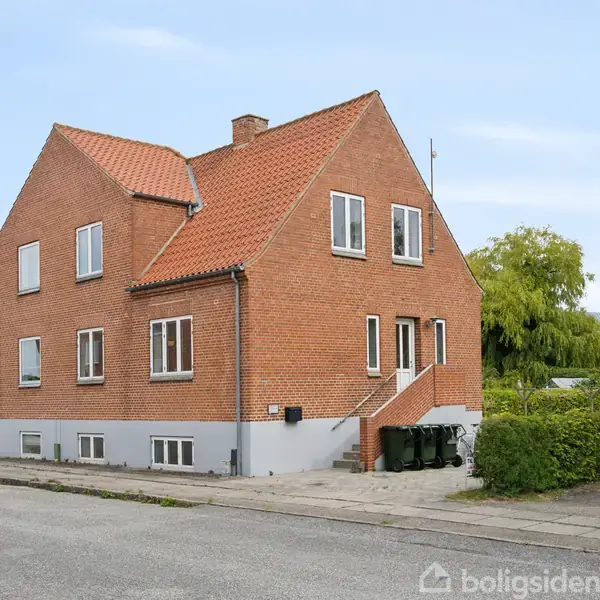 Rødt murstenshus med rødt tegltag og hvidrammede vinduer. Foran huset er en trappe med gelænder, og tre skraldespande står ved siden af. Omgivet af hække og andre huse i nabolaget.