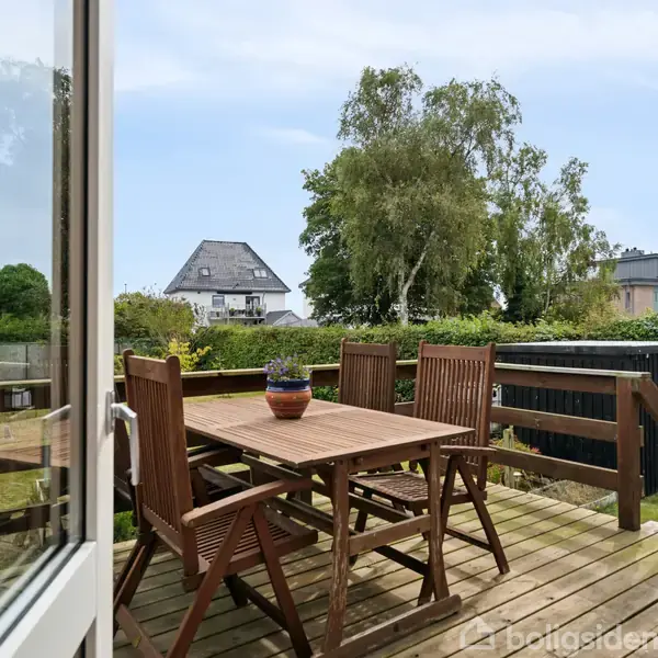 Et træbord med fire stole står på en terrasse med udsigt til en have. En krukke står på bordet. I baggrunden ses et hus og træer.