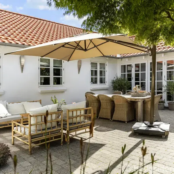 Udendørs gårdhave med møbleret siddeområde under stor parasol. Rattanmøbler og bord er placeret på flisebelægning foran hvidt hus med rødt tegltag. Grønne planter omgiver området.