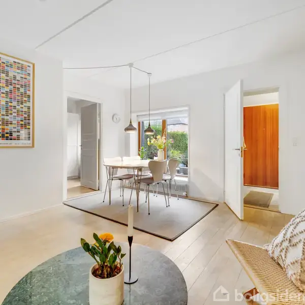 Et lyst og moderne stue med et rundt sofabord, vase med blomster og en hvid væg med et stort, farverigt billede. Spisebordet ses i baggrunden ved et vindue.