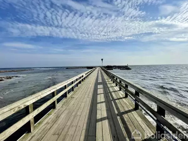 Træmolen strækker sig ud mod havet under en blå, let skyet himmel. Havet omgiver den smalle trækonstruktion, der fører mod en fjerntliggende mole yderst i billedet.