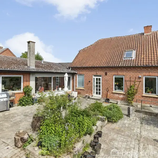Rødt murstenshus med saddeltag omkranser en flisebelagt gårdhave. Grill står til venstre. Grønne planter og små træstubbe dekorerer området under en blå himmel med spredte skyer.