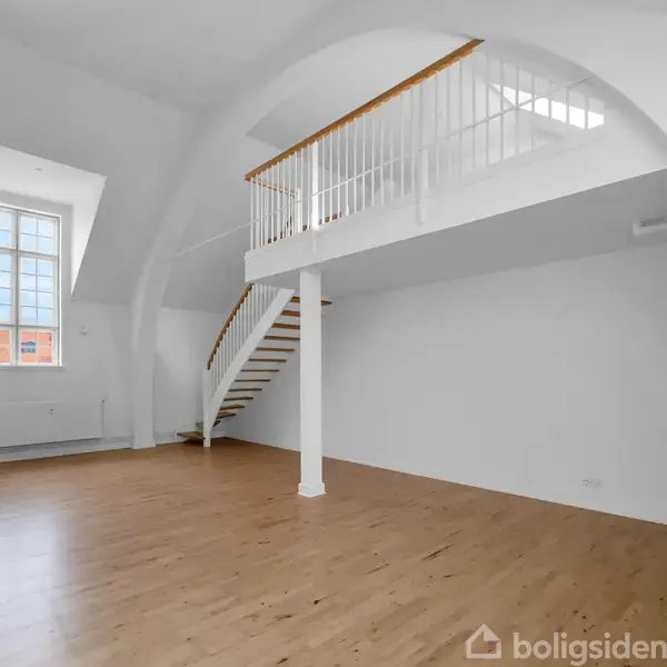 En stor, lys stue har et loft med buede hvide vægge. En trappe fører op til en indvendig altan, og der er et stort vindue i baggrunden.