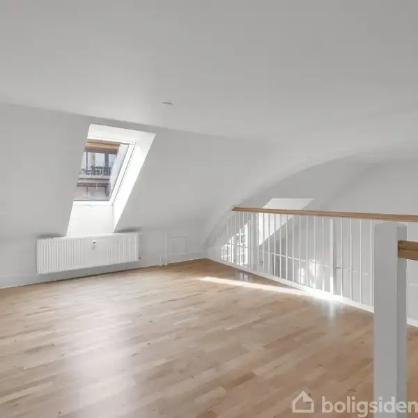 Et tomt rum med skråt loft og et ovenlysvindue, der slipper lys ind. Rummet har et lyst trægulv, hvidmalede vægge og en trappe med hvidt gelænder mod højre.