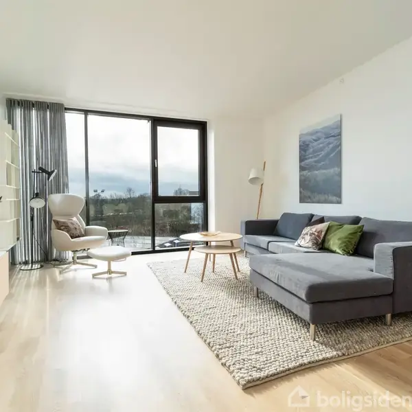 Grå sofa står i en lys stue med trægulv. To små borde foran sofaen. Store vinduer giver udsigt til udendørsområdet. Plante og stol i rummets hjørne tilføjer charme.