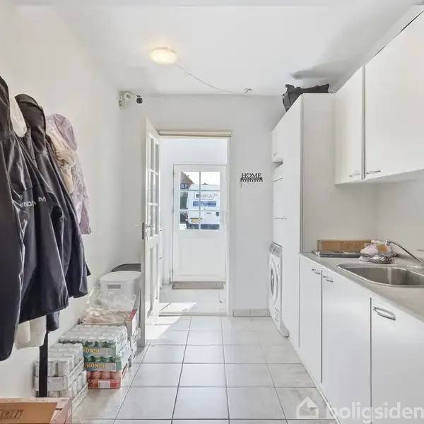 Jakker hænger på en vægkrog i et bryggers. Vaskemaskine er indbygget i hvide skabe. Der er en dør til et lyst rum i baggrunden.