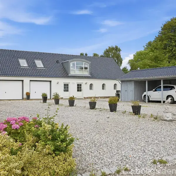 En stor hvid bygning med sort tag har flere vinduer og garageporte. En bil holder parkeret under en carport. Omgivet af en grusbelagt gårdsplads med blomsterkrukker og buske.