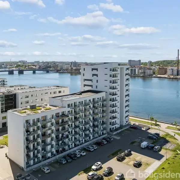 Et lejlighedskompleks med altaner står stille. Omkringliggende biler er parkeret på asfalten. I baggrunden ses en flod, en bro og flere bygninger i en byudsigt.