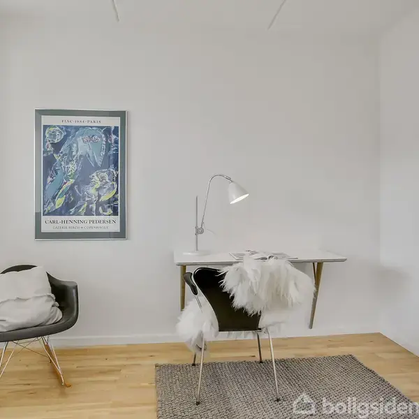 Et skrivebord med en lampe står i et lyst rum. En stol med et fluffy tæppe er ved siden af. På væggen hænger en plakat: "Carl-Henning Pedersen".