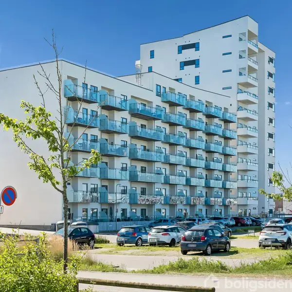 En hvid, moderne lejlighedsbygning med flere altaner står i et byområde. Biler er parkeret foran bygningen, og der er grønne områder og klare himmel i baggrunden.