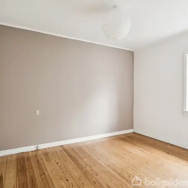 Værelse med trægulv og hvid væg, brun accentvæg til venstre. Hængende hvid lampe i loftet. Vindue til højre viser træer udenfor. Radiator under vinduet.