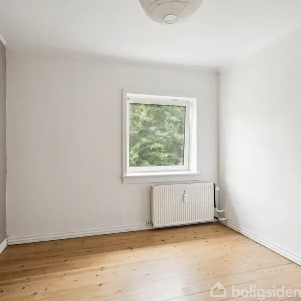 Et tomt rum med træplankegulv og hvide vægge. Et vindue viser grønne træer udenfor. En radiator er placeret under vinduet. Rummet er lyst og neutralt indrettet.