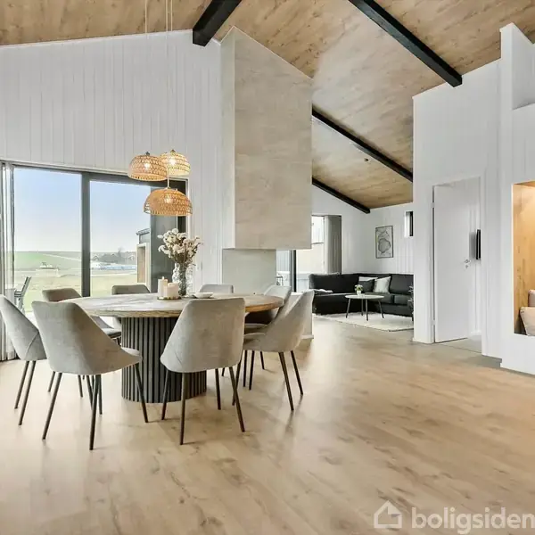En spisestue med rundt bord og otte grå stole, pyntet med en vase. Store vinduer giver udsigt over et grønt landskab. Træloft og skrå vægge skaber en indbydende atmosfære.