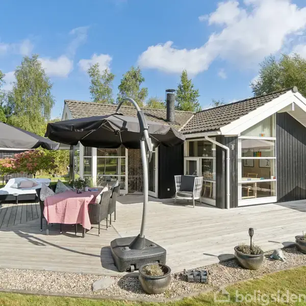 Et moderne sommerhus med en træterrasse, hvor der står havemøbler og parasoller. Baggrunden har grøn beplantning og en klar blå himmel, hvilket giver en sommerlig atmosfære.