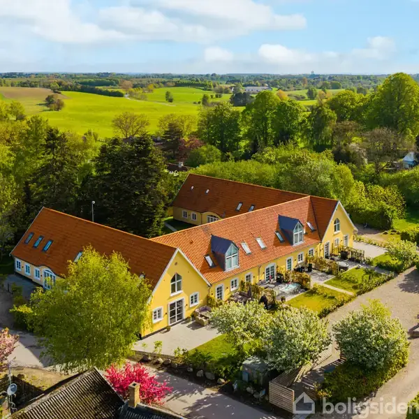 Et gult bondehus med rødt tegltag ligger i en frodig landskabskontekst omgivet af grønne marker og træer. Foran huset er der en velplejet have med blomstrende træer.
