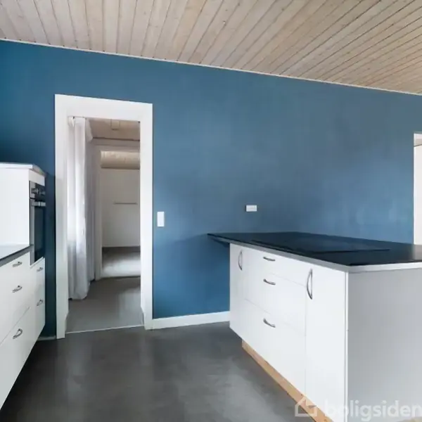 Et hvidt køkken med en kogeø med sort bordplade. Køkkenet har hvide skabe, en ovn og en blå væg som baggrund. Døre fører til tilstødende rum.