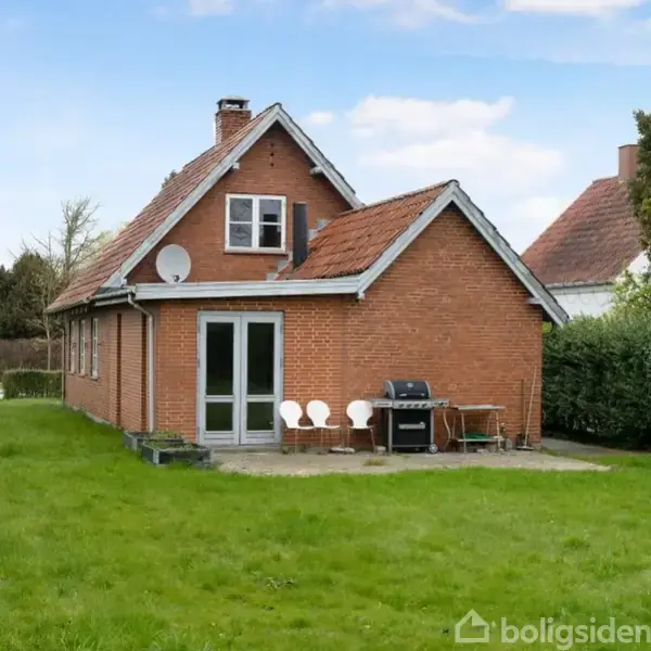 Rødt murstenshus med saddeltag står i en velplejet have. Et sæt dobbeltdøre åbner mod en terrasse med tre stole og en grill. Skyfri himmel i baggrunden.