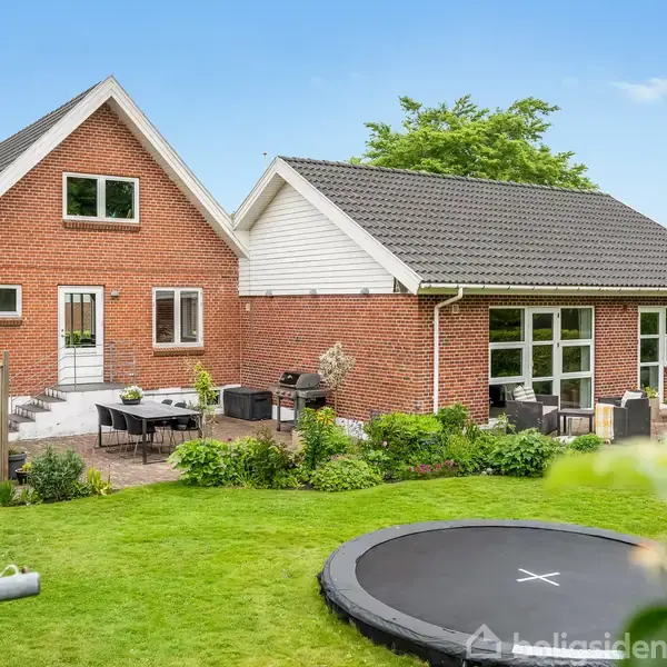 Rødt murstenshus med stort sort tag; udestue med store vinduer. Terrasse med havemøbler ses foran huset. Omkringliggende have har græsplæne og trampolin. Solrig dag med blå himmel.