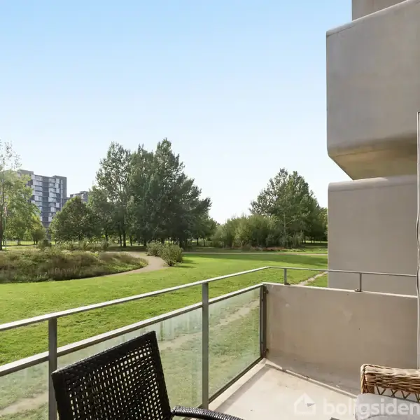 Balkon med glasrækværk og en stol. Åben dør leder til en lejlighed. Udsigt over en grøn park med stier og træer, samt højhuse i baggrunden.