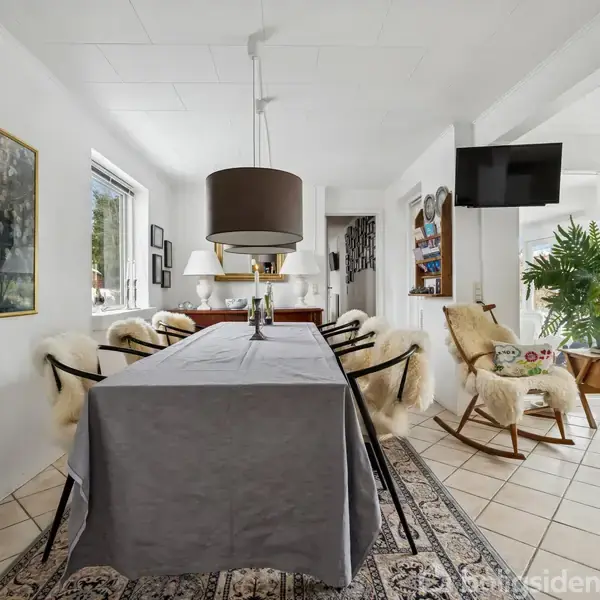 Et langbord med gråt tæppe er omringet af stole med lyse skind. En stor lampe hænger over bordet. Rummet har lyse vægge, store vinduer og en lejlighedsfjerner i baggrunden.