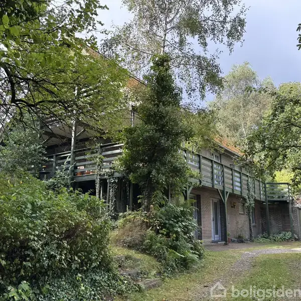 Et hus i mursten står omgivet af tæt bevoksning og store træer. Huset har en stor, omkransende træterrasse, der hæver sig over jorden. Atmosfæren er fredfyldt og naturrig.